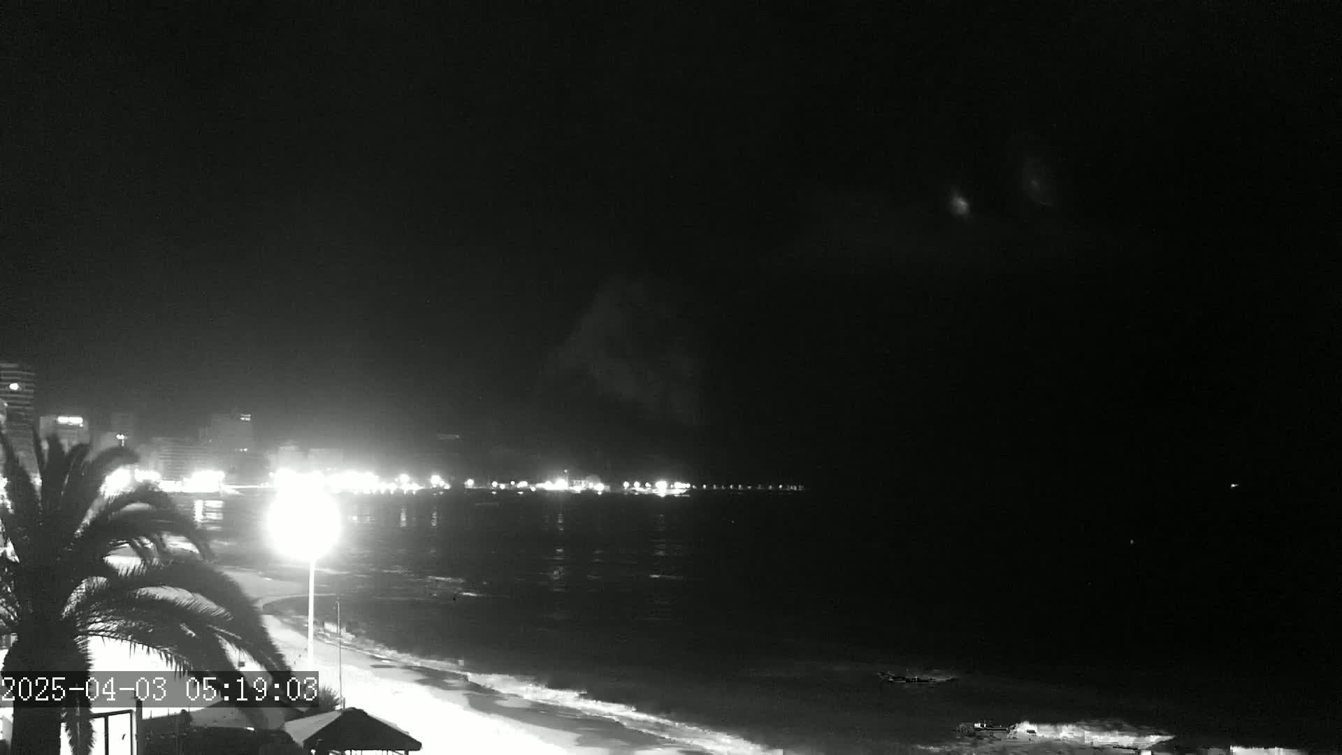 A nighttime view of a beach with a city skyline in the background under a mostly clear sky.