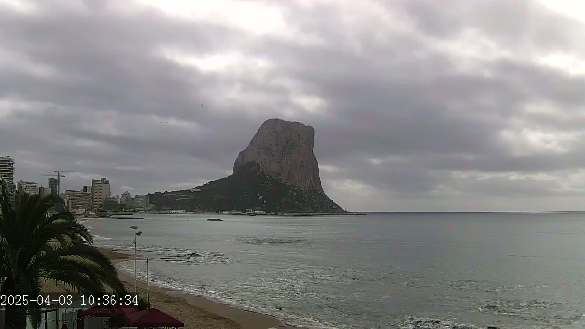 A large, imposing rock formation is seen across a calm bay from a beach with a city skyline in the background under an overcast sky.