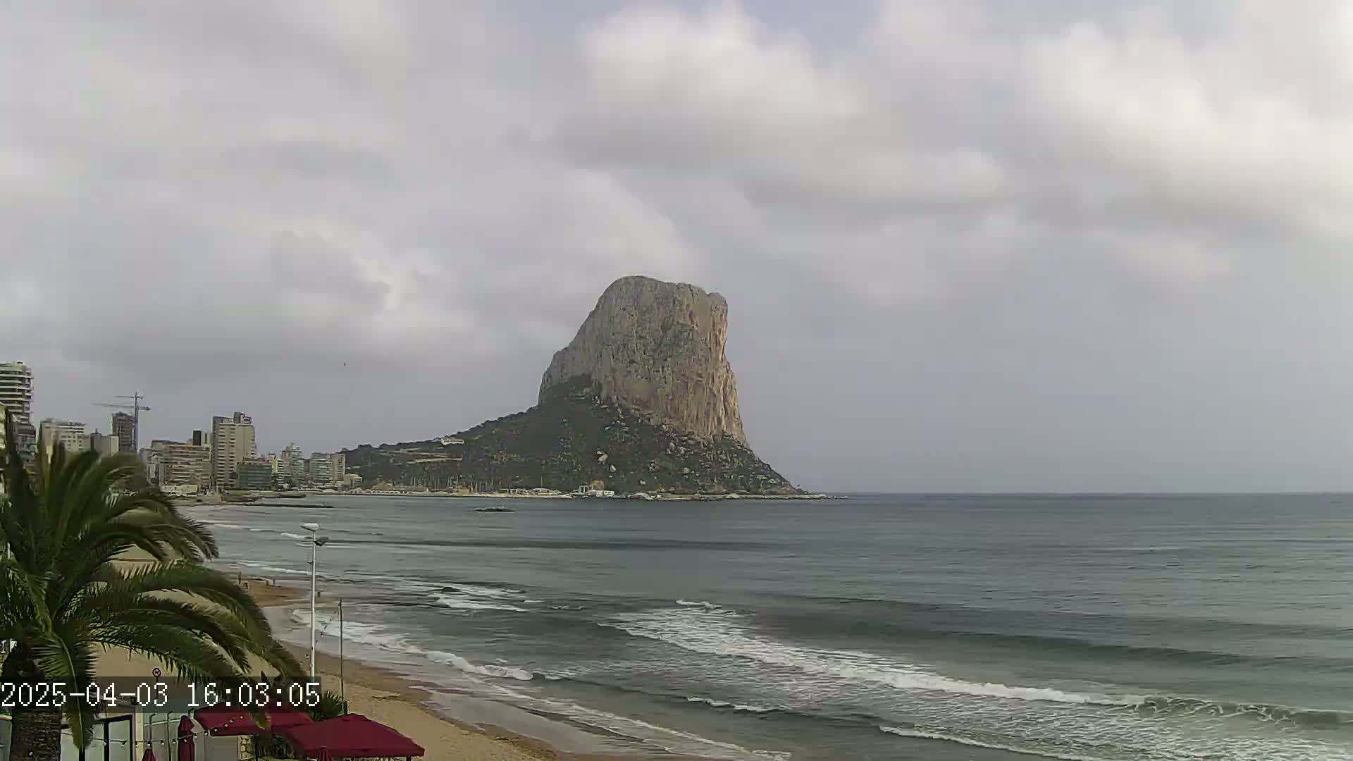 A large, rocky island rises from the sea under a cloudy sky, with a sandy beach and buildings visible in the foreground.