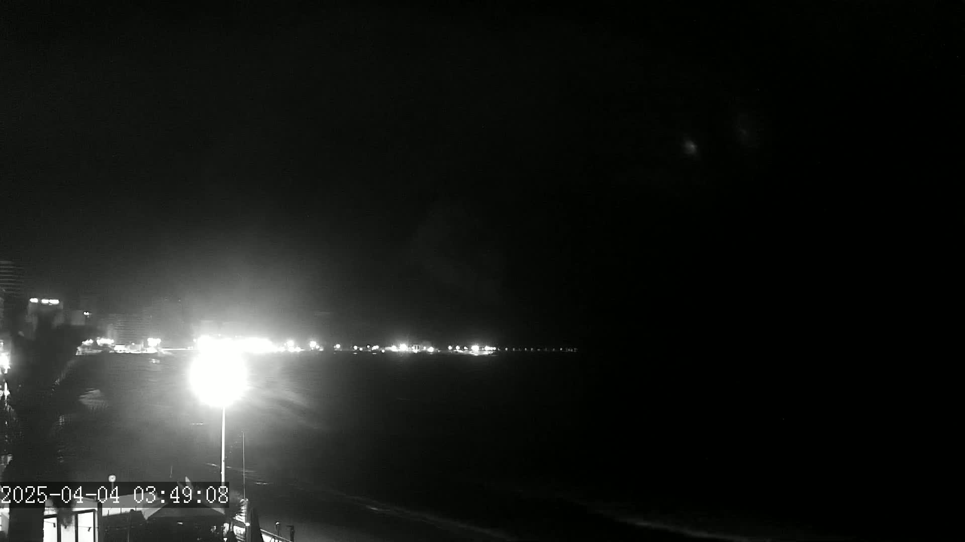 A nighttime, low-light view shows a dimly lit shoreline with buildings and lights in the distance, under a dark, clear sky.