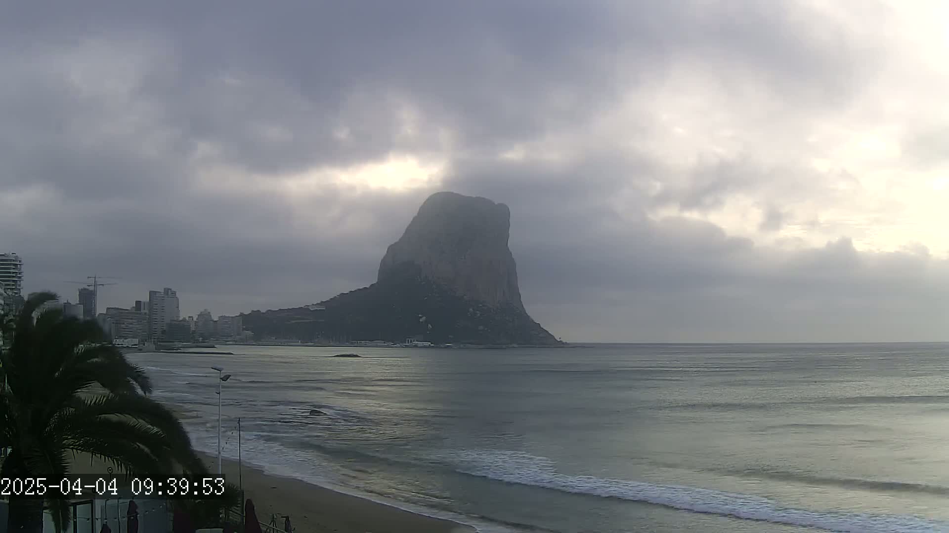 A large, dark rock formation rises from the ocean under a mostly cloudy sky, with a city visible in the background on the left, and a sandy beach in the foreground.