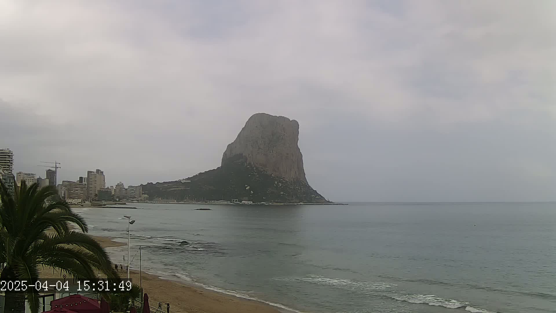 A large, rocky island is visible across a calm, grey sea from a beach with buildings and palm trees in the foreground under an overcast sky.