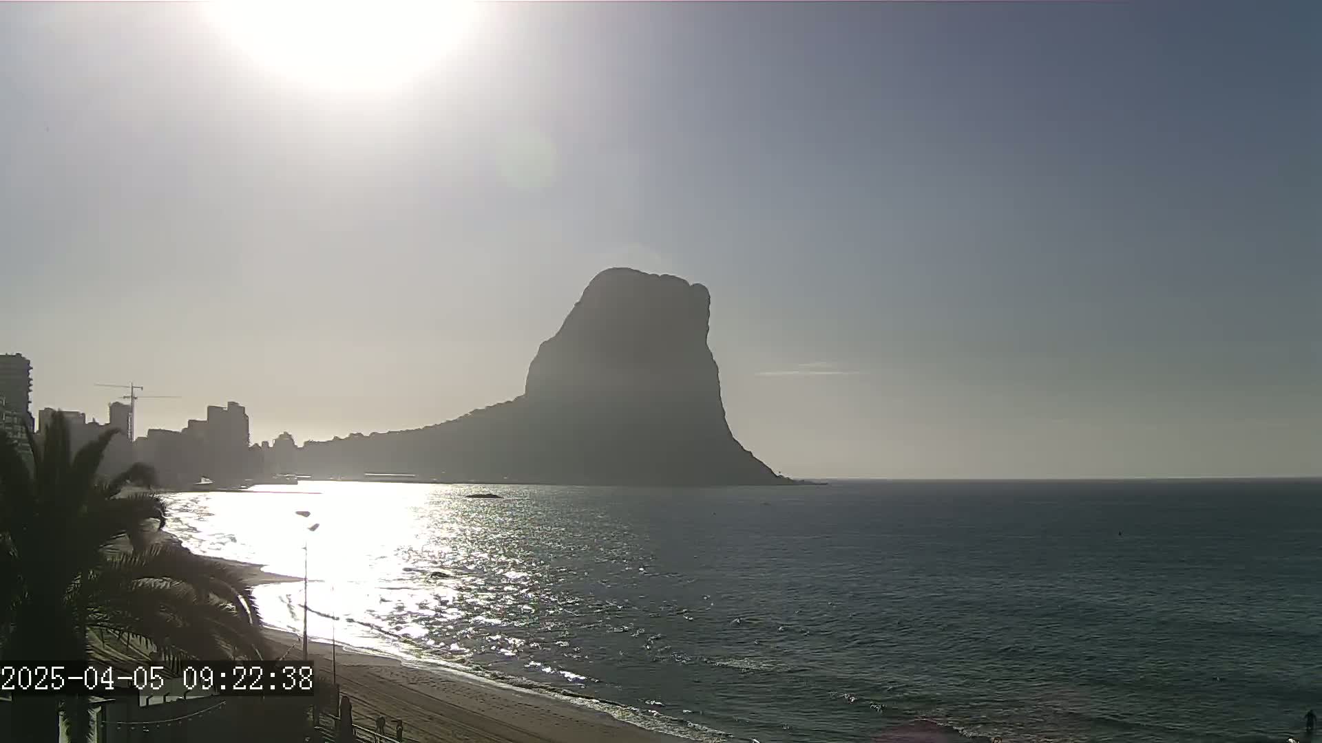 A large, dark, solitary hill stands prominently in the distance across a calm, sunlit ocean, with a sandy beach and buildings visible in the foreground on a clear day.