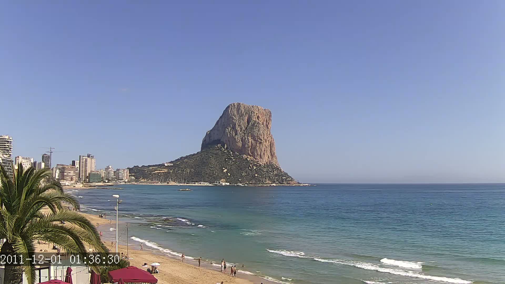 A sandy beach with calm ocean waves and a few people, overlooks a city skyline and a large, rocky, mountainous island under a clear blue sky.