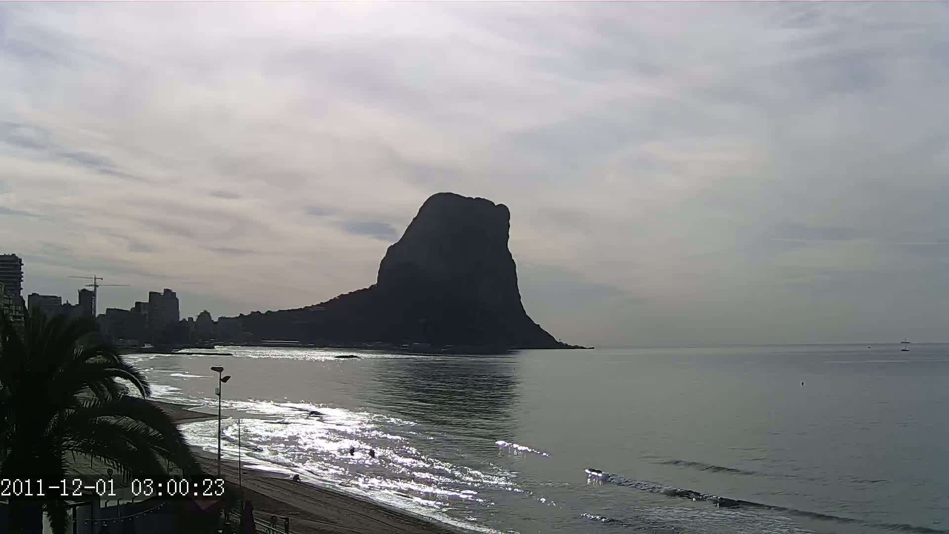 A large, dark mountain rises from the calm sea near a beach with gentle waves, under a cloudy sky.