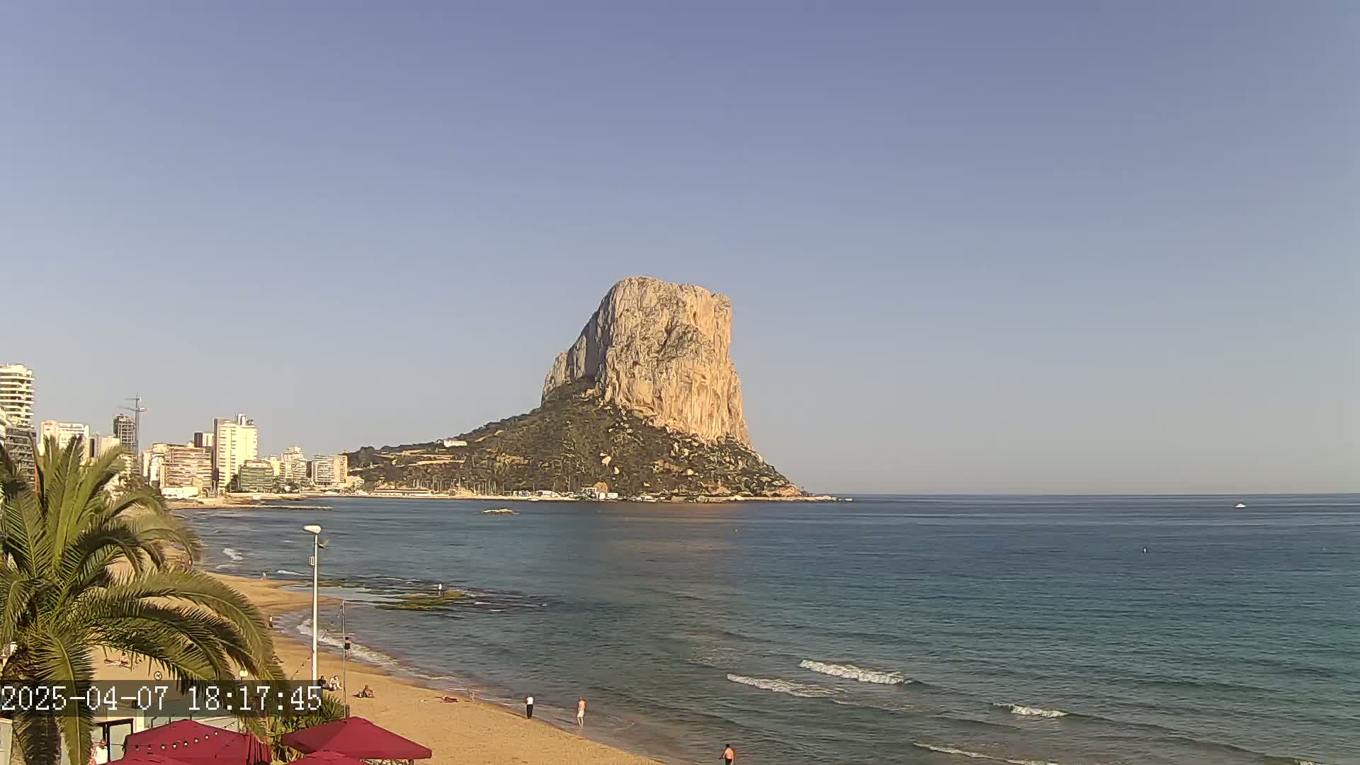 A large, light-brown rock formation rises from the sea, visible from a sandy beach with several people on it, under a clear blue sky.