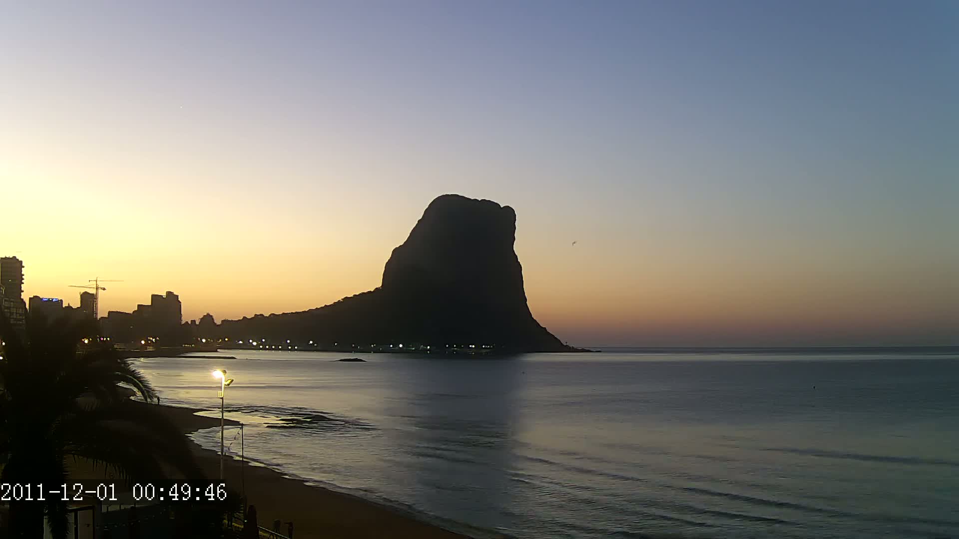 A large, dark silhouette of a rock formation rises from the sea at sunrise, overlooking a city skyline and beach.