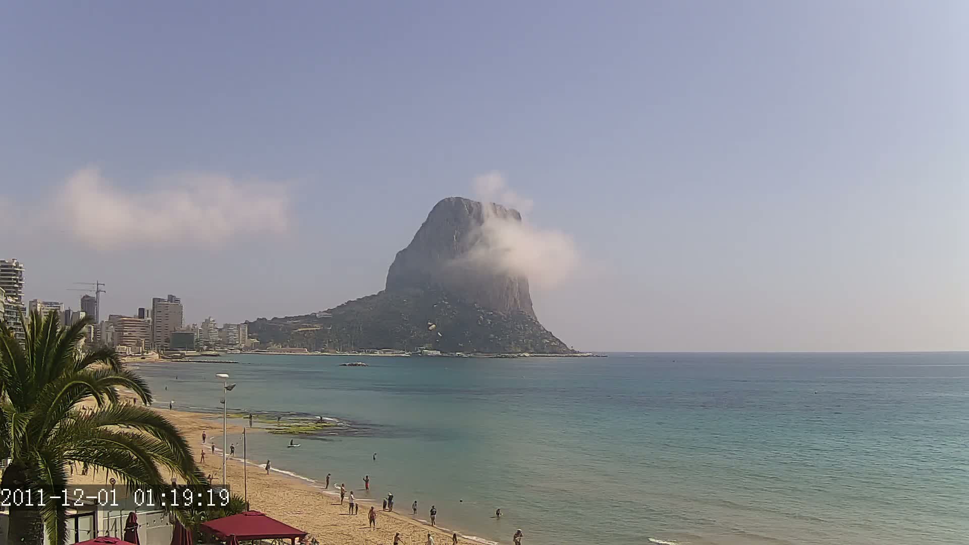 A sandy beach with scattered people stretches along a calm, teal sea towards a large, rocky, cloud-covered mountain under a mostly clear sky.