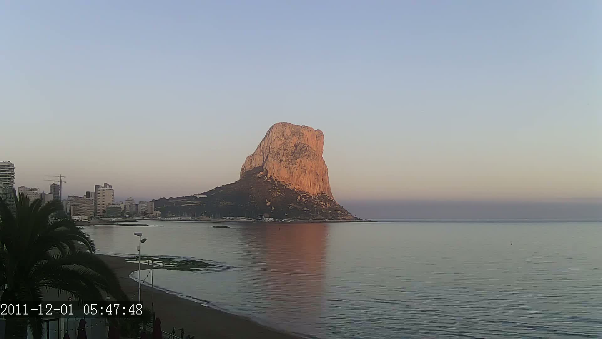 A large, light-brown rock formation rises from the calm, reflective waters of a bay at sunrise, with a city skyline visible to the left.