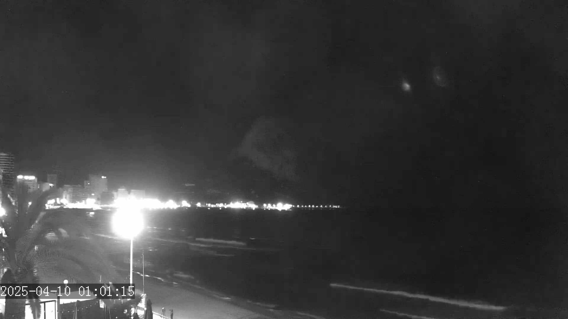 A nighttime view of a beach with a city skyline in the background, under a dark, cloudy sky.