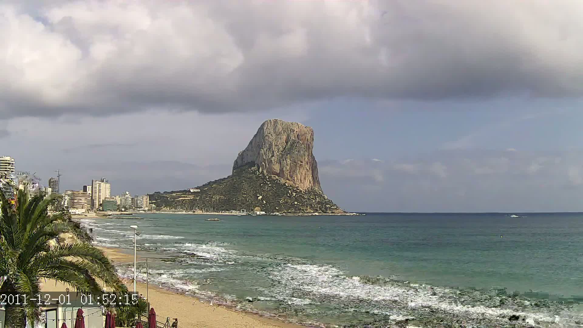 A large, rocky island stands prominently in the distance across a wavy ocean under a partly cloudy sky, with a sandy beach and buildings visible along the coastline.