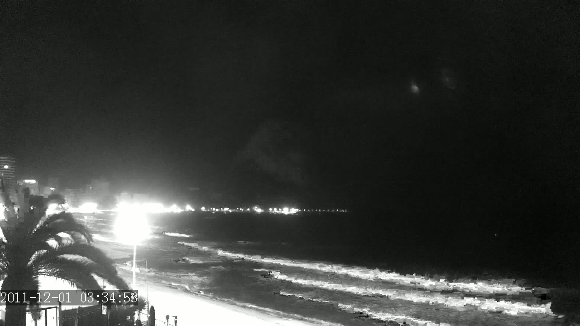 A nighttime view of a beach with waves gently lapping the shore, illuminated by lights from buildings along the coast under a dark sky.