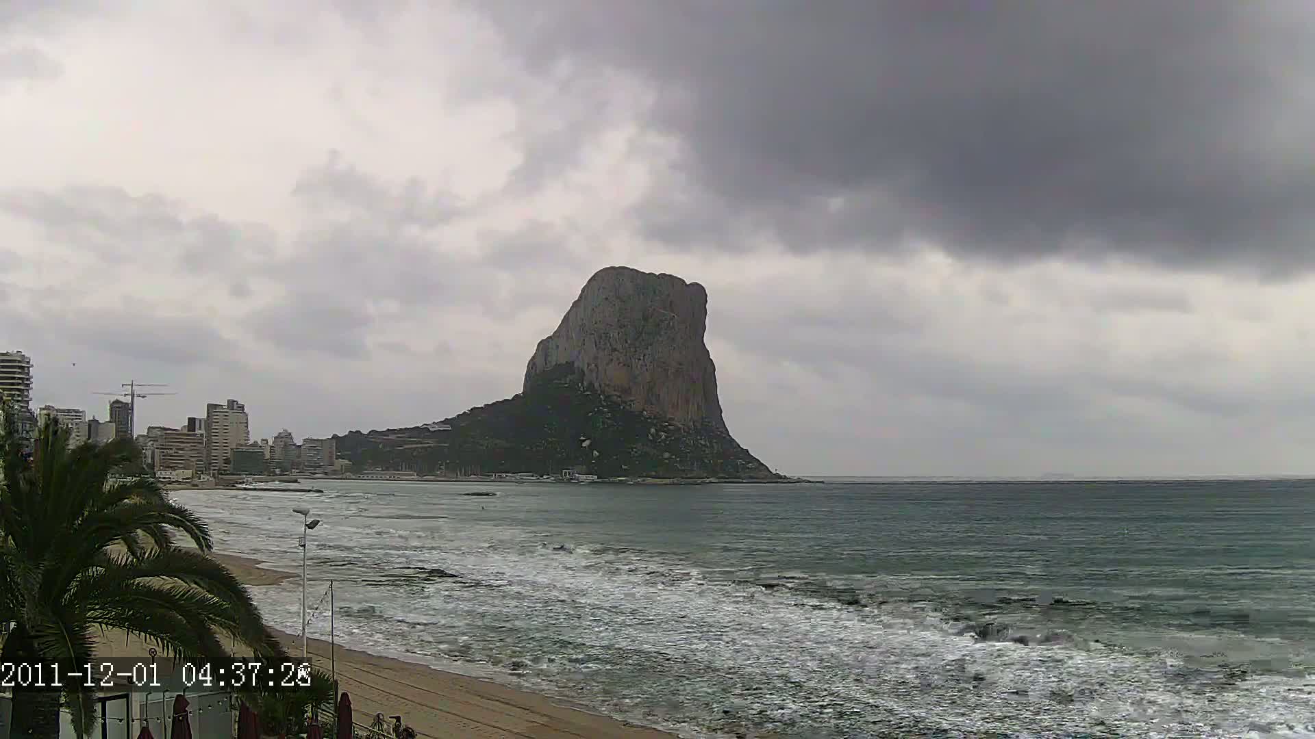 A large, dark rock formation rises from the sea under a cloudy sky, with a city skyline and sandy beach visible in the foreground.