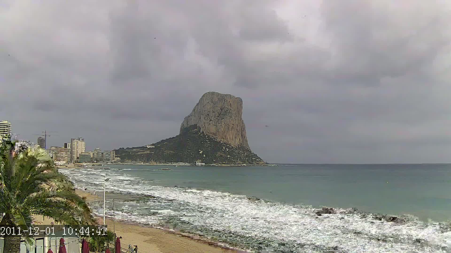 A large, imposing rock formation rises from the sea on an overcast day, with waves breaking on a sandy beach in front of a cityscape.
