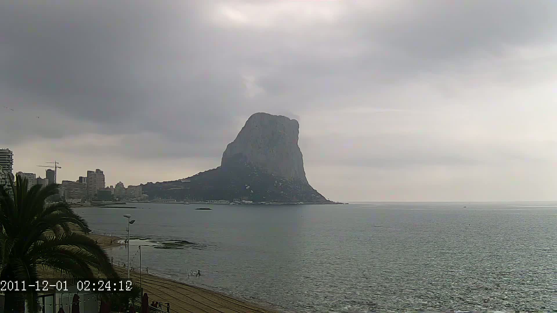 A large, rocky outcrop rises from the sea under an overcast sky, with a beach and buildings visible in the foreground.