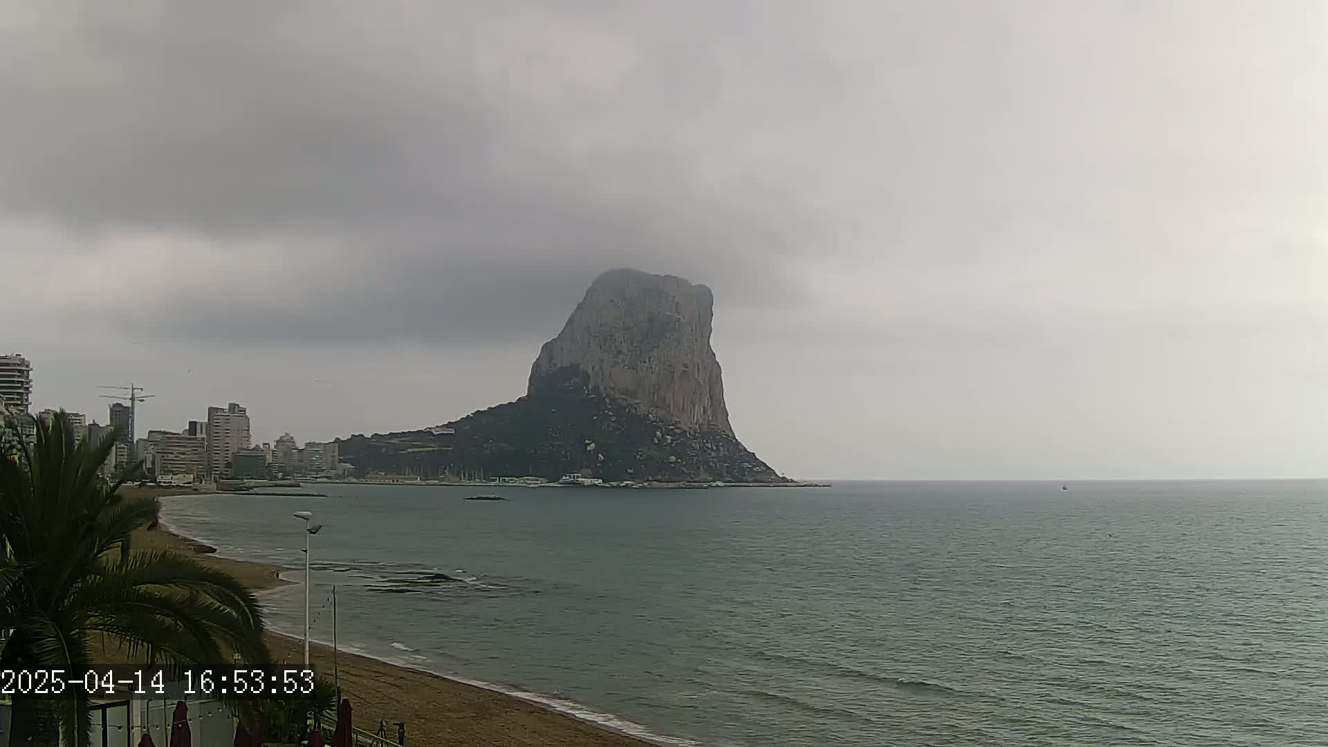 A large, dark rock formation rises from the sea under an overcast sky, visible from a beach with buildings and palm trees in the foreground.
