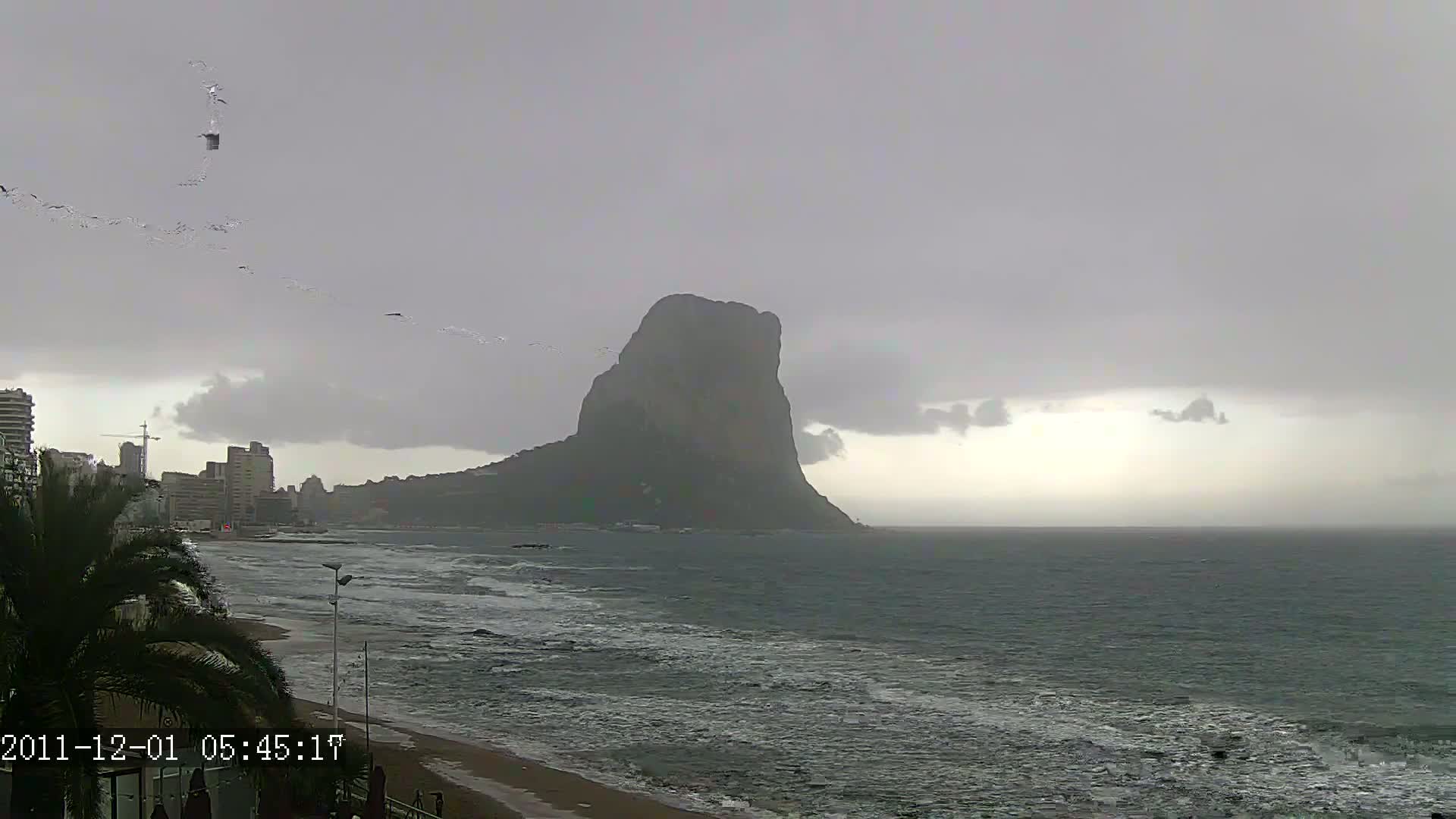 A large, dark rock formation rises from the sea under an overcast sky, with choppy waves on the shore and a city skyline visible in the distance.