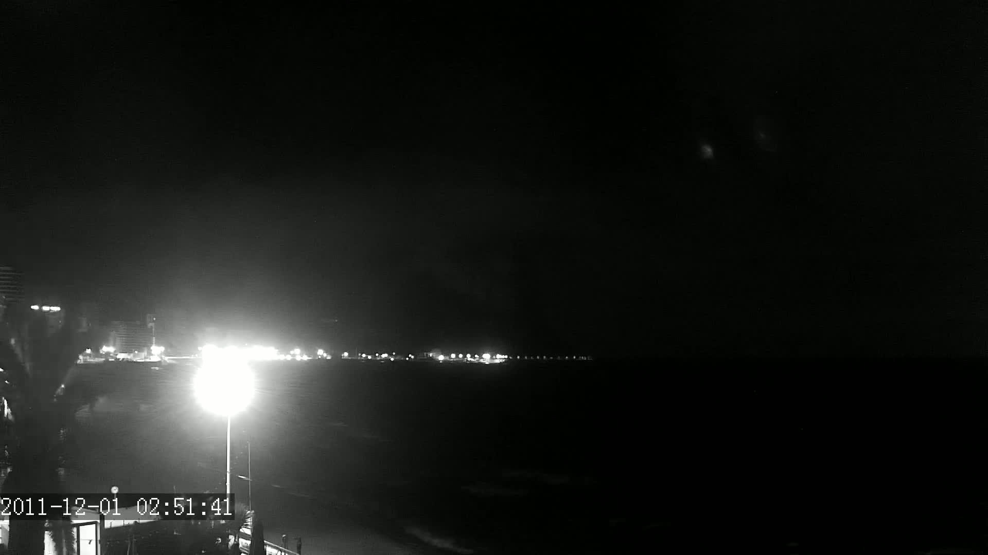 A nighttime, low-light view shows a dark beach with a distant line of city lights under a clear, dark sky.