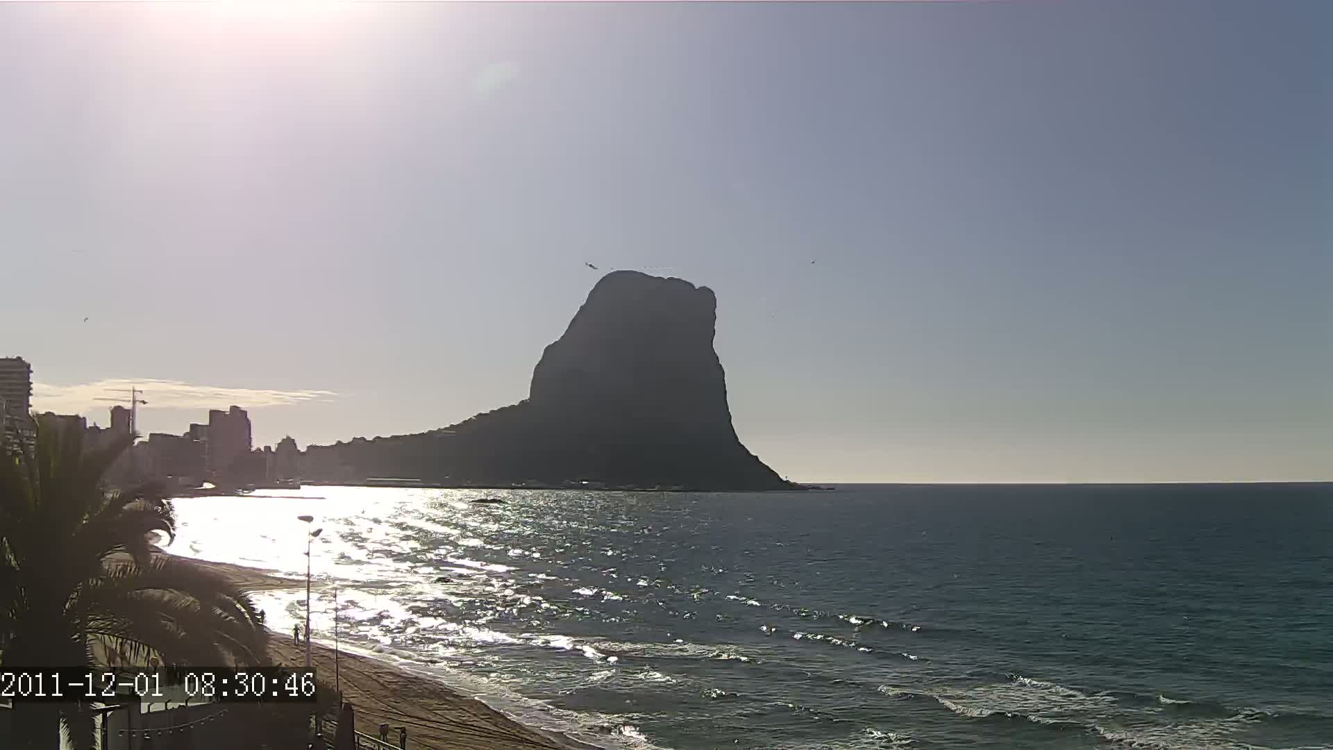 A large, dark mountain rises from the sea on a sunny day, with a sandy beach and buildings visible in the foreground.