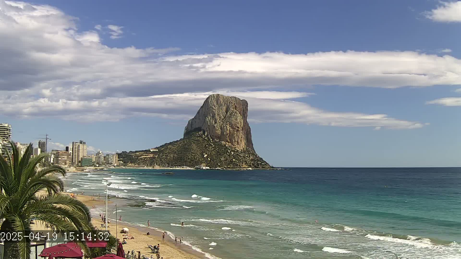 A sandy beach with people on it fronts a calm, turquoise ocean, with a large, rocky island visible in the distance under a partly cloudy sky.