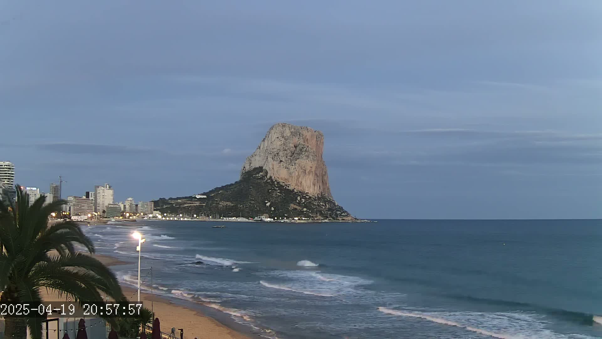 A large, rocky cliff stands prominently in the distance across a calm, twilight ocean, with a beach and cityscape visible in the foreground.