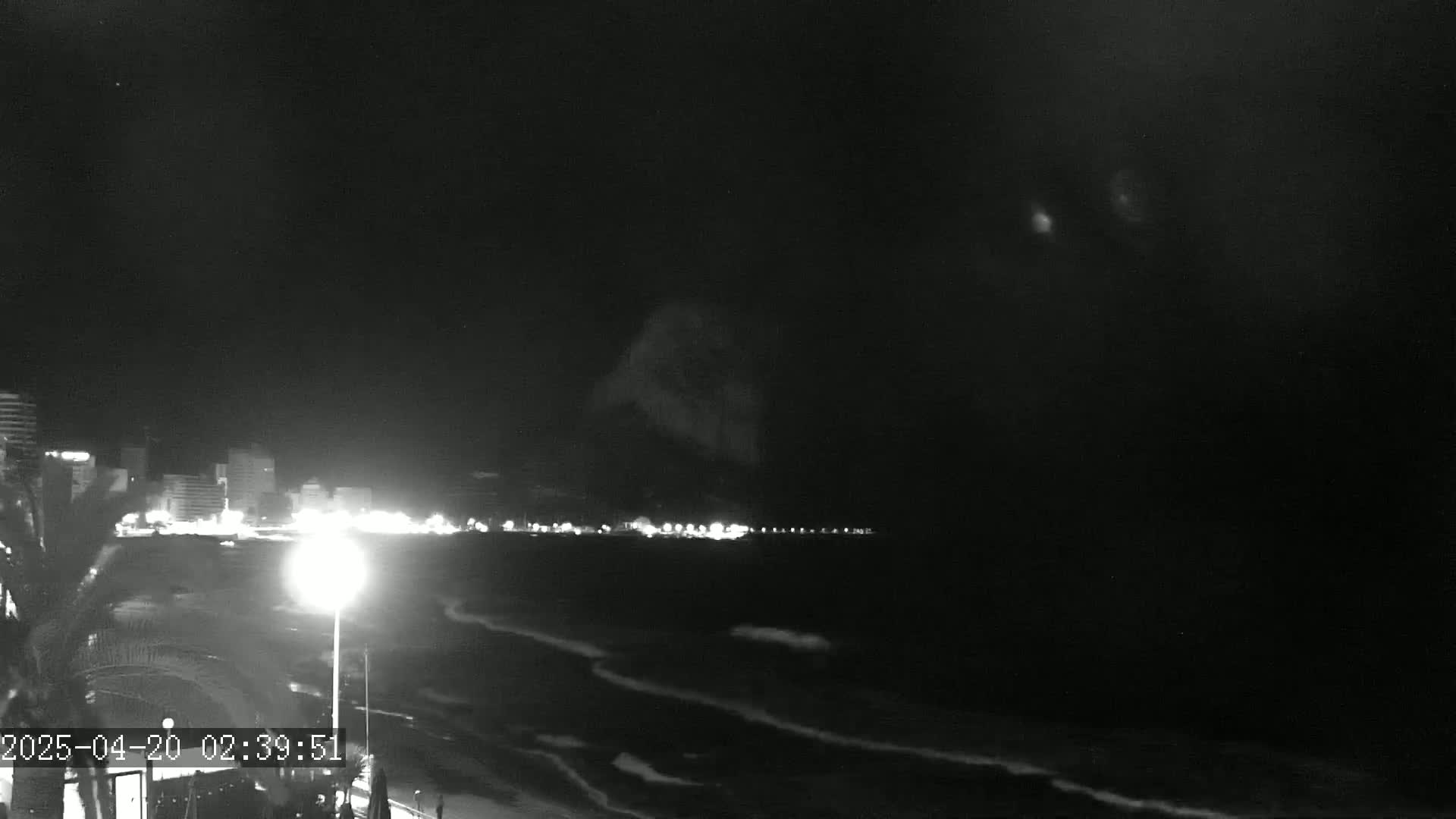 A nighttime, black and white view shows a dimly lit beach with gentle waves, a row of buildings across the water, and a dark sky.