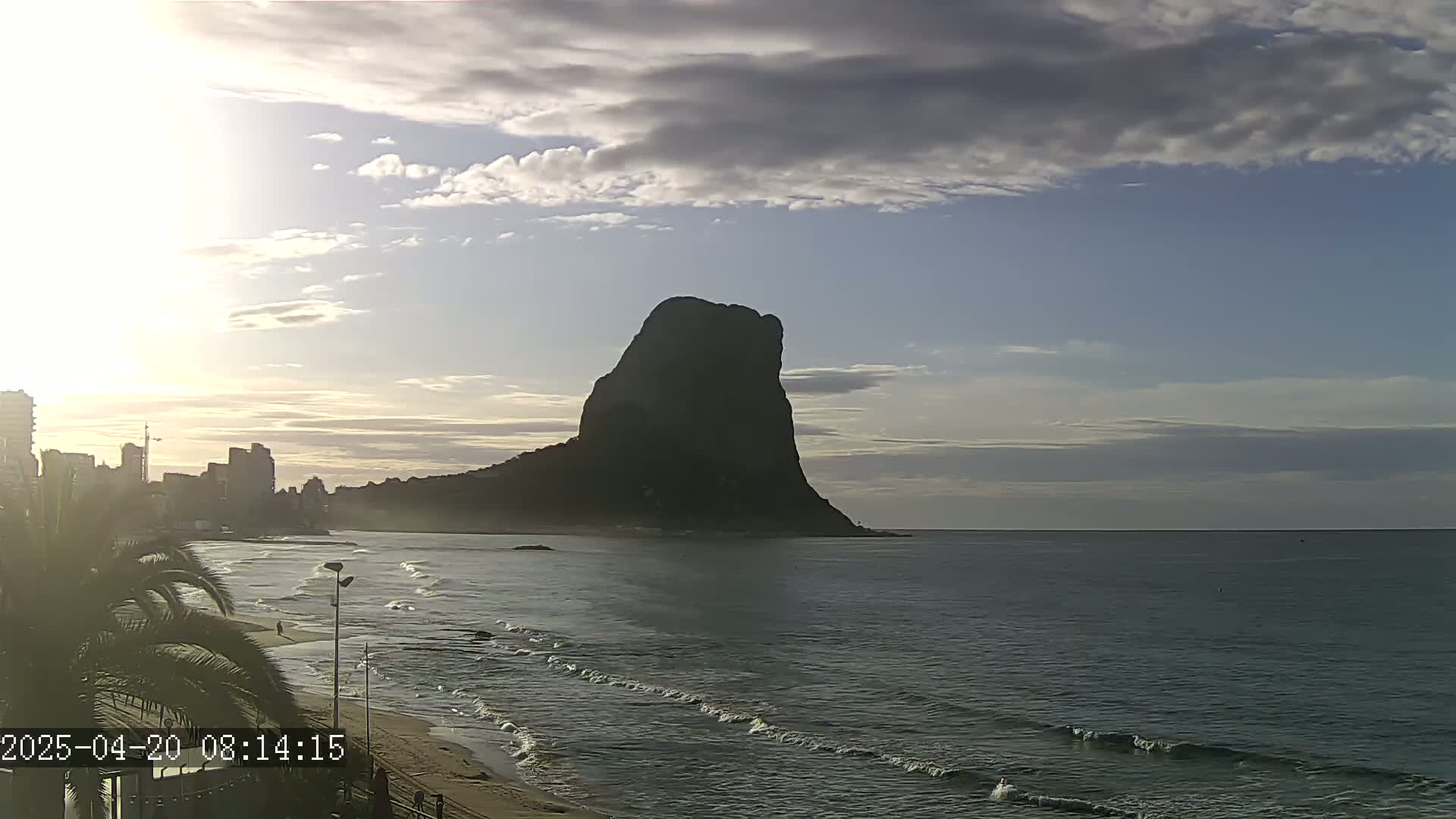A large, dark rock formation rises from the ocean under a partly cloudy sky, with a beach and buildings visible in the foreground.
