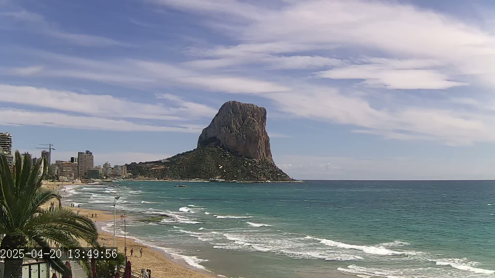 A large, rocky island rises from a teal ocean, with a sandy beach and buildings visible on the shore under a partly cloudy sky.
