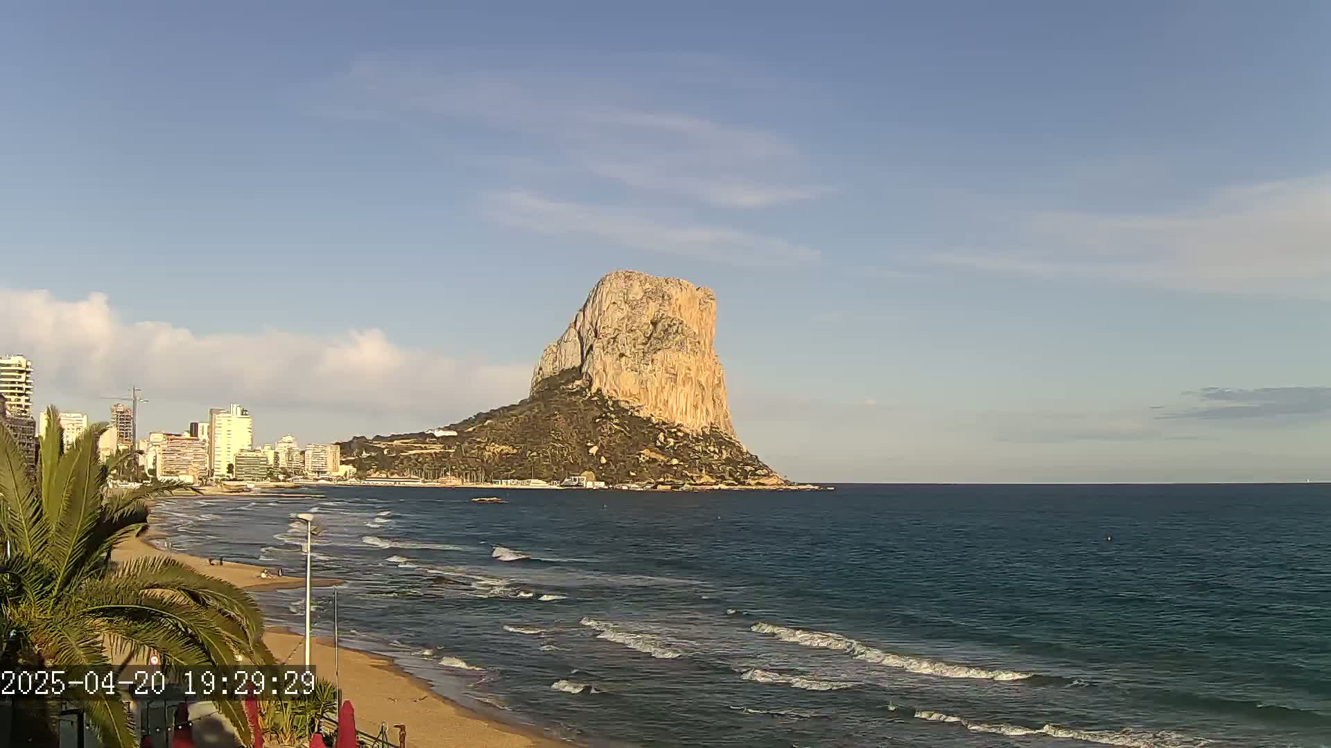 A large, light-brown rock formation rises from the sea near a sandy beach and a city skyline under a mostly clear sky.