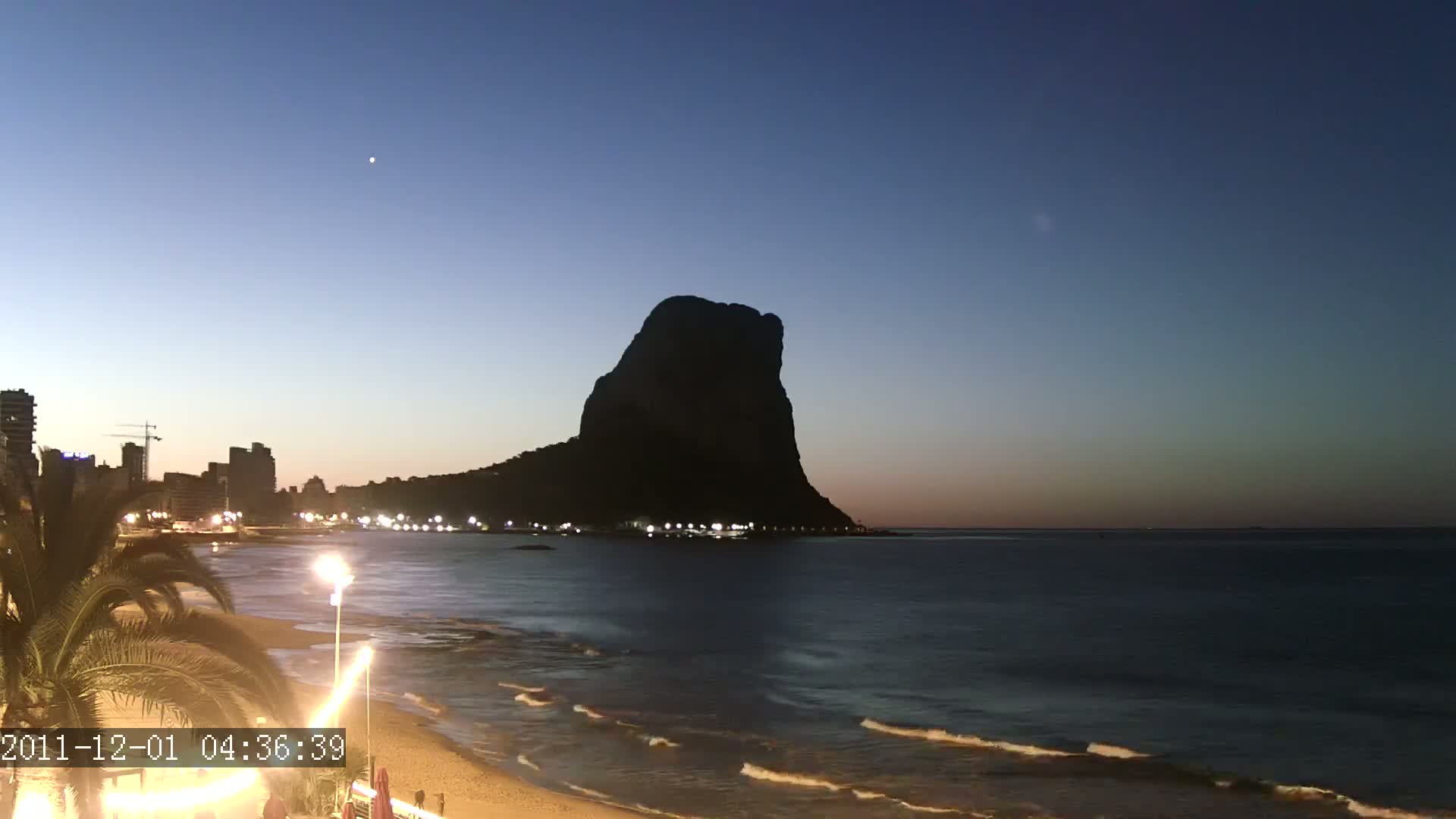 A dark, silhouetted mountain rises above a beach at twilight, with calm waves lapping the shore under a clear, dark-blue sky.