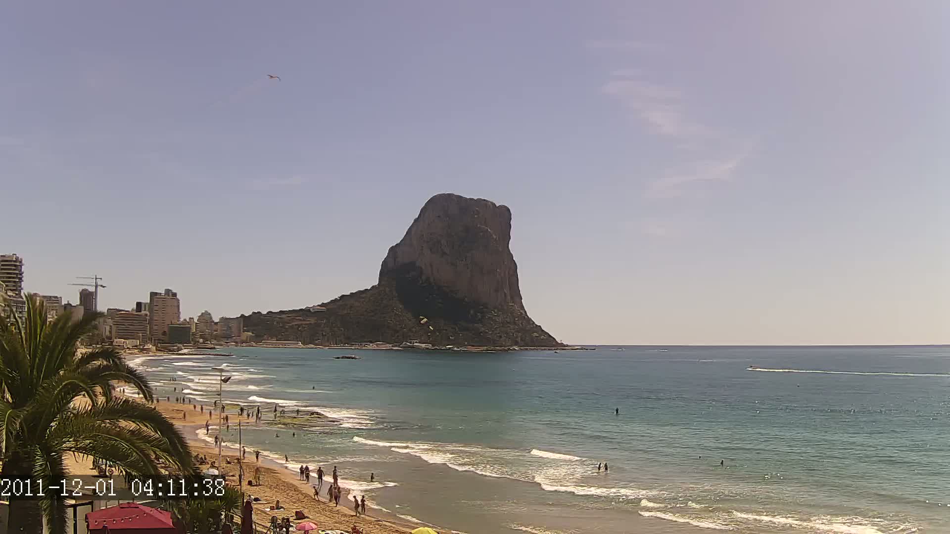 A sandy beach with people sunbathing and swimming is visible in front of a large, dark rock formation rising from the sea under a clear, bright sky.