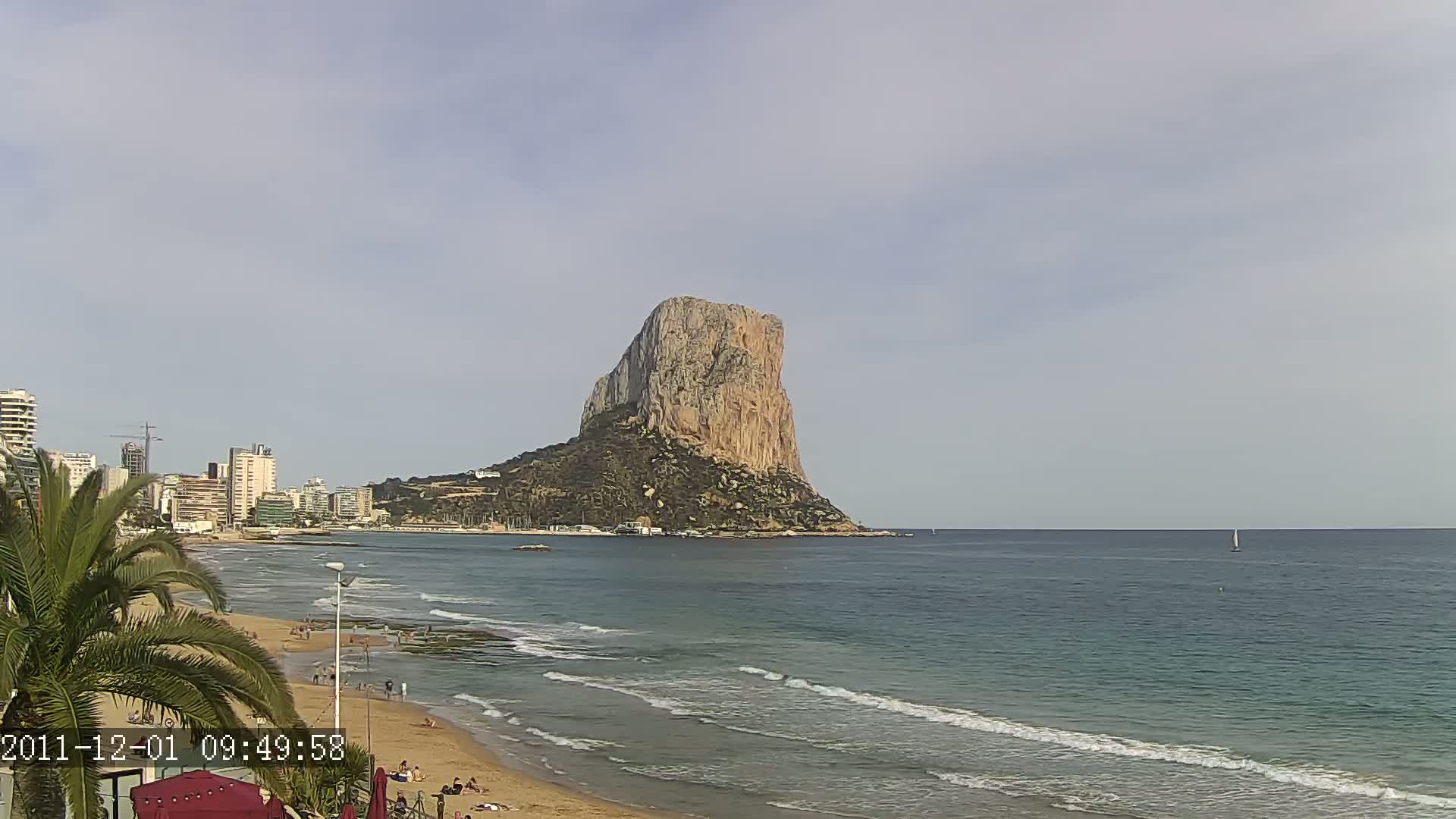 A large, tan rock formation rises from the sea near a sandy beach with gentle waves under a mostly clear sky.