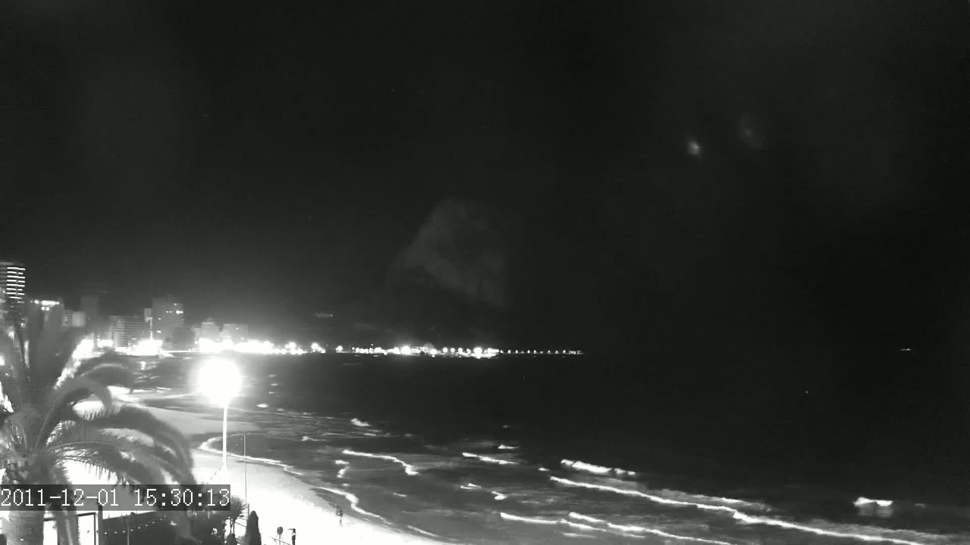 A nighttime beach scene shows gently rolling waves under a dark sky with a line of city lights in the distance.