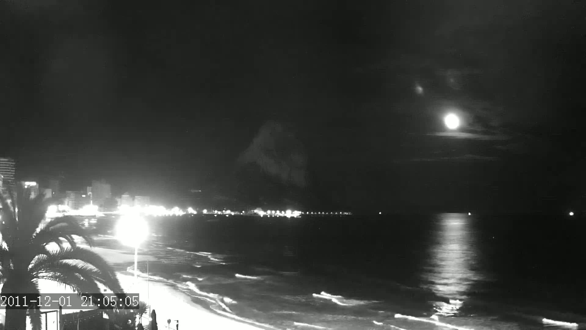 A bright moon illuminates a nighttime beach scene with calm waves lapping the shore, and a city skyline visible in the distance.