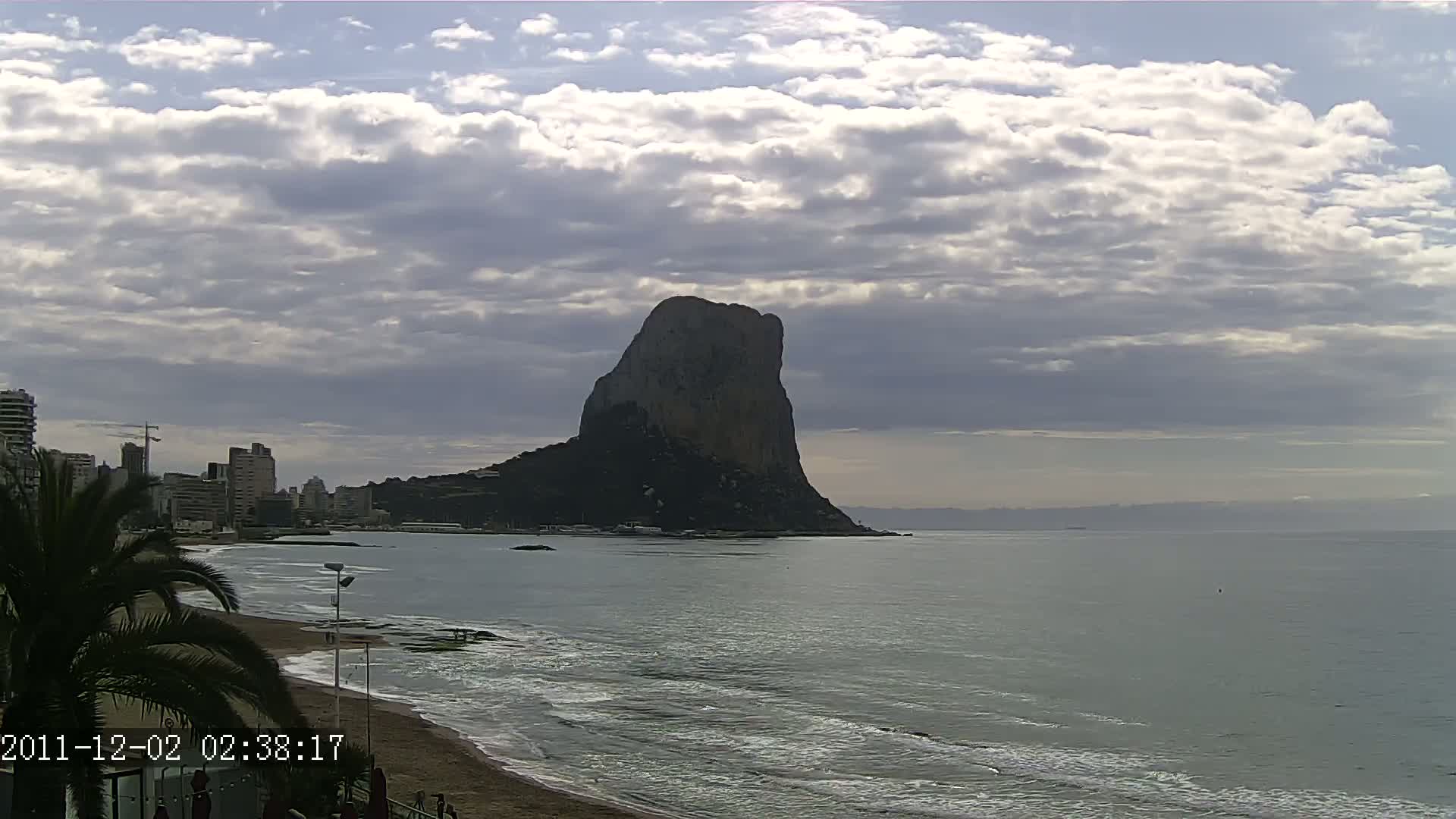 A large, dark rock formation rises from the sea under a partly cloudy sky, with a beach and buildings visible in the foreground.