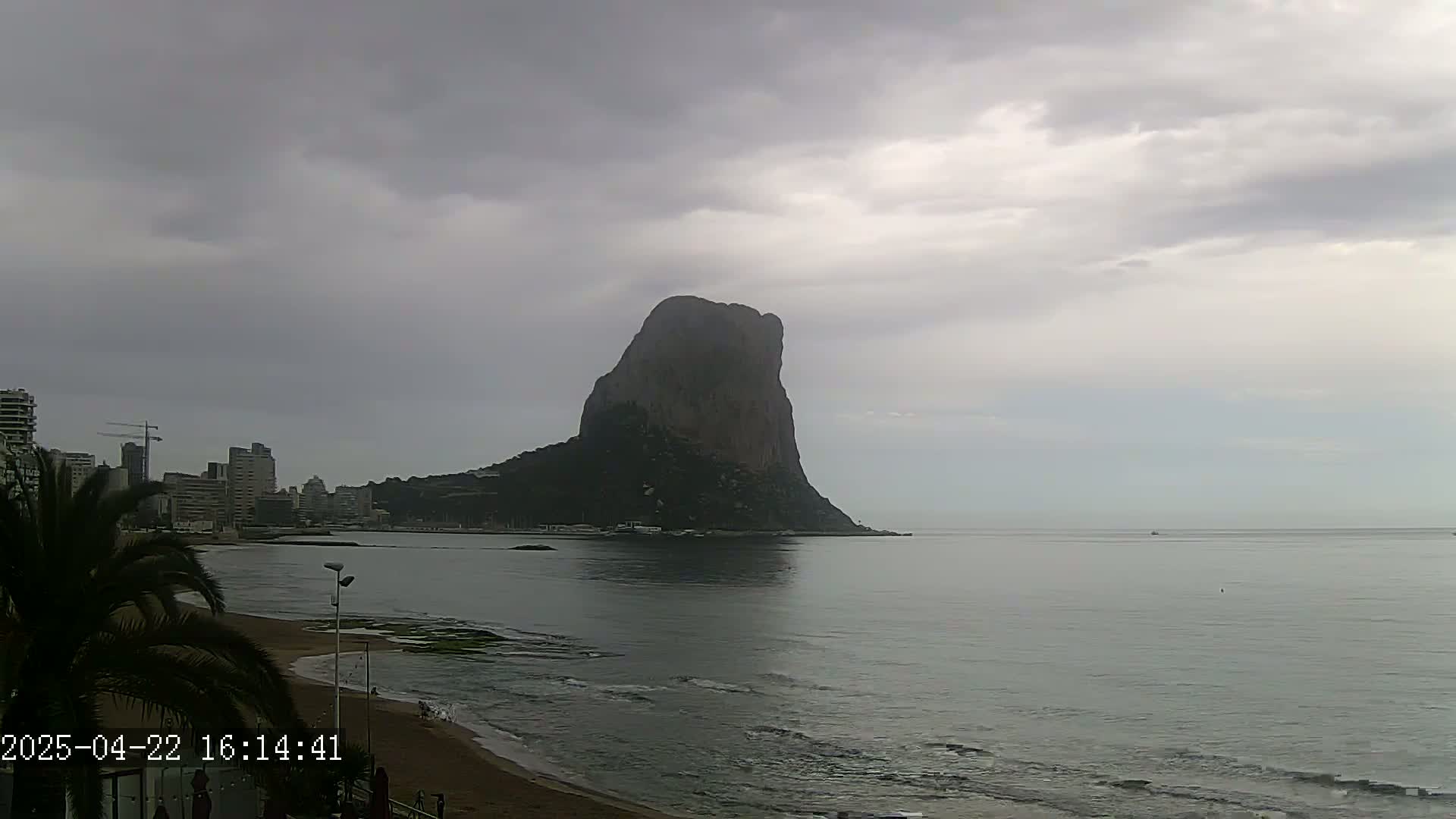 A large, dark rock formation rises from the sea under a cloudy sky, with a beach and cityscape visible to the left.