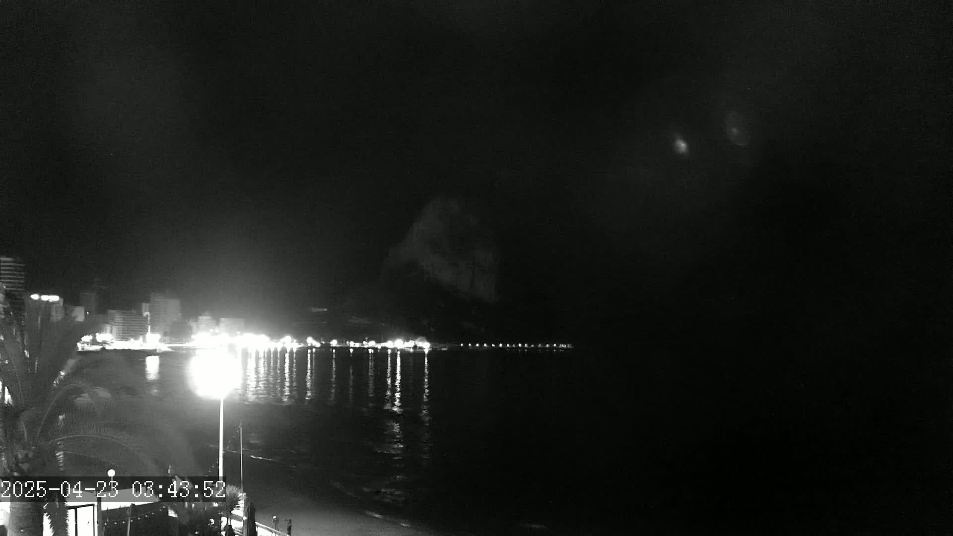 A nighttime view of a dark, calm ocean reflecting the lights of a city skyline and a hill in the distance under a cloudy sky.