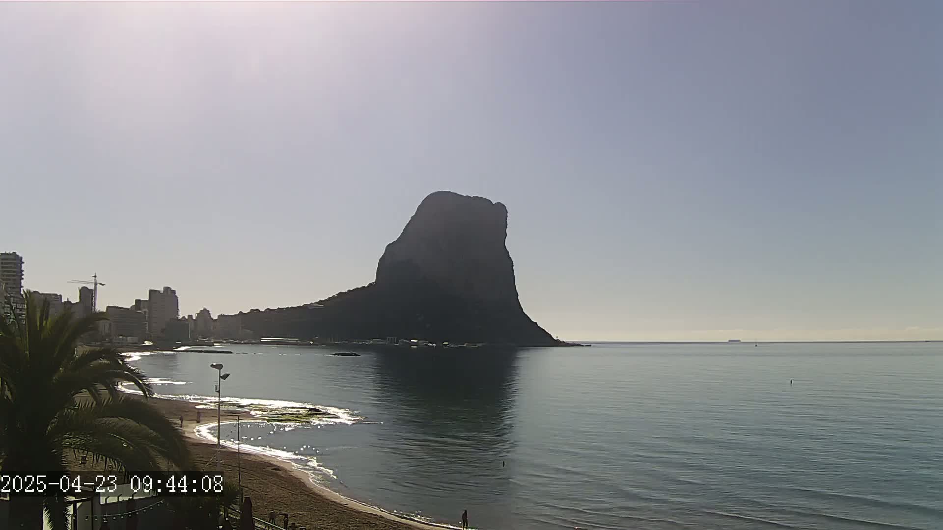 A large, dark rock formation rises from the calm, light-blue sea near a beach with buildings and palm trees under a bright, clear sky.