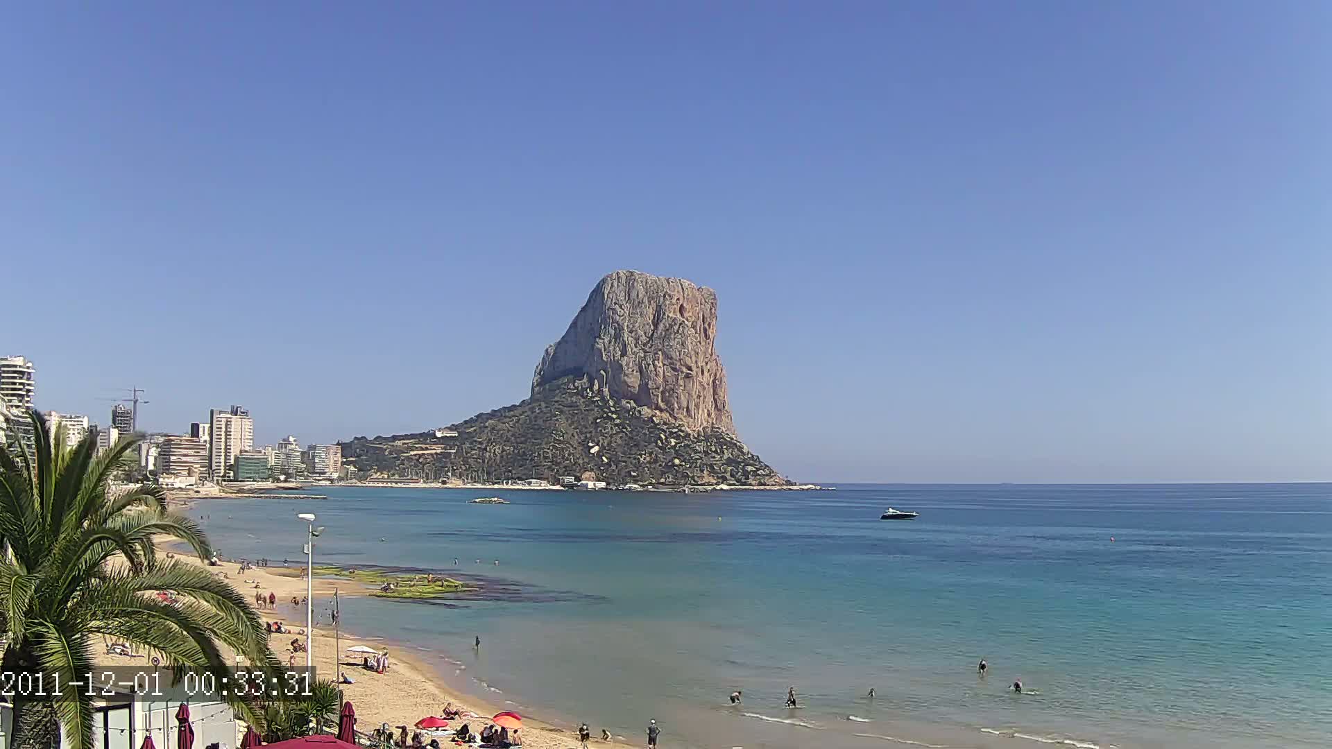 A sandy beach with people swimming and sunbathing stretches before a calm, blue sea, with a large, rocky outcrop rising from the water in the background under a clear, blue sky.