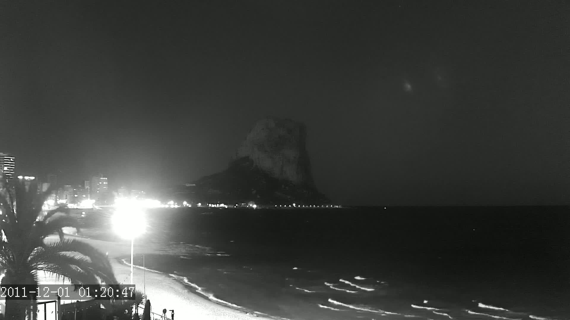 A large, dark rock formation stands in the distance across a gently lapping sea at night, with a dimly lit beach and buildings in the foreground.