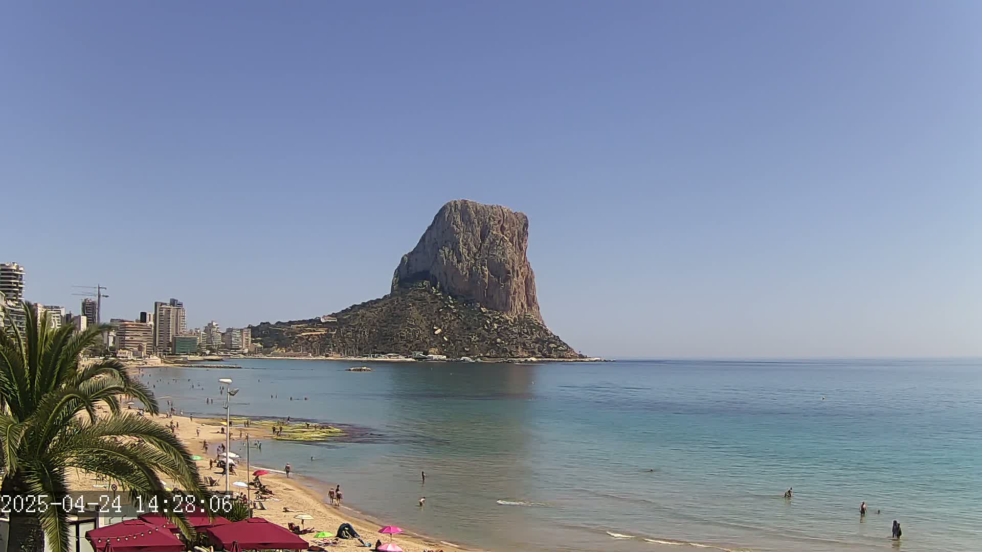 A sandy beach with people swimming and sunbathing stretches out before a large, rocky mountain that rises from the calm, clear ocean under a bright blue sky.