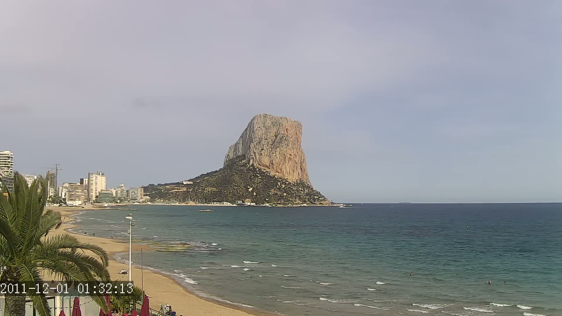 A large, tan rock formation rises from the sea near a sandy beach and beachfront buildings under an overcast sky.