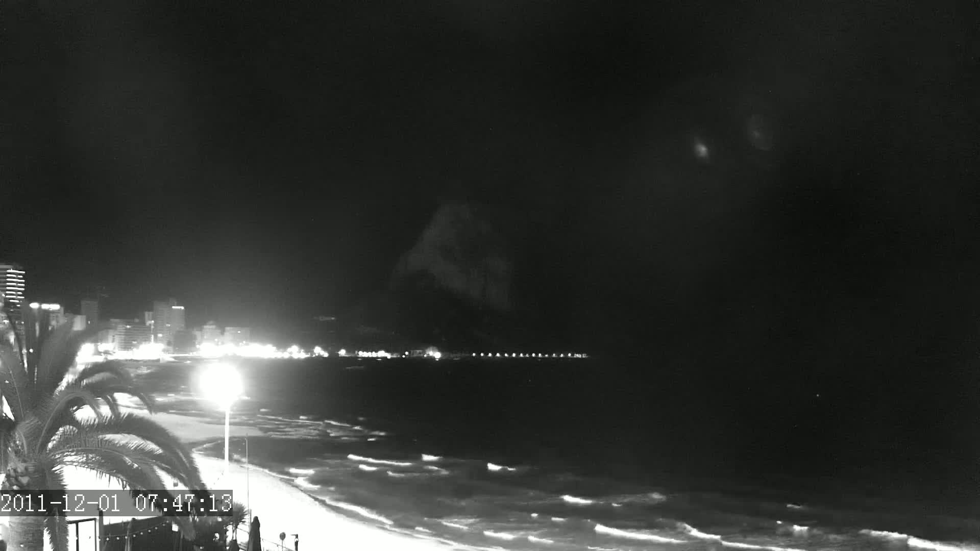 A nighttime beach scene shows a city skyline across a gently waving ocean under a dark sky.