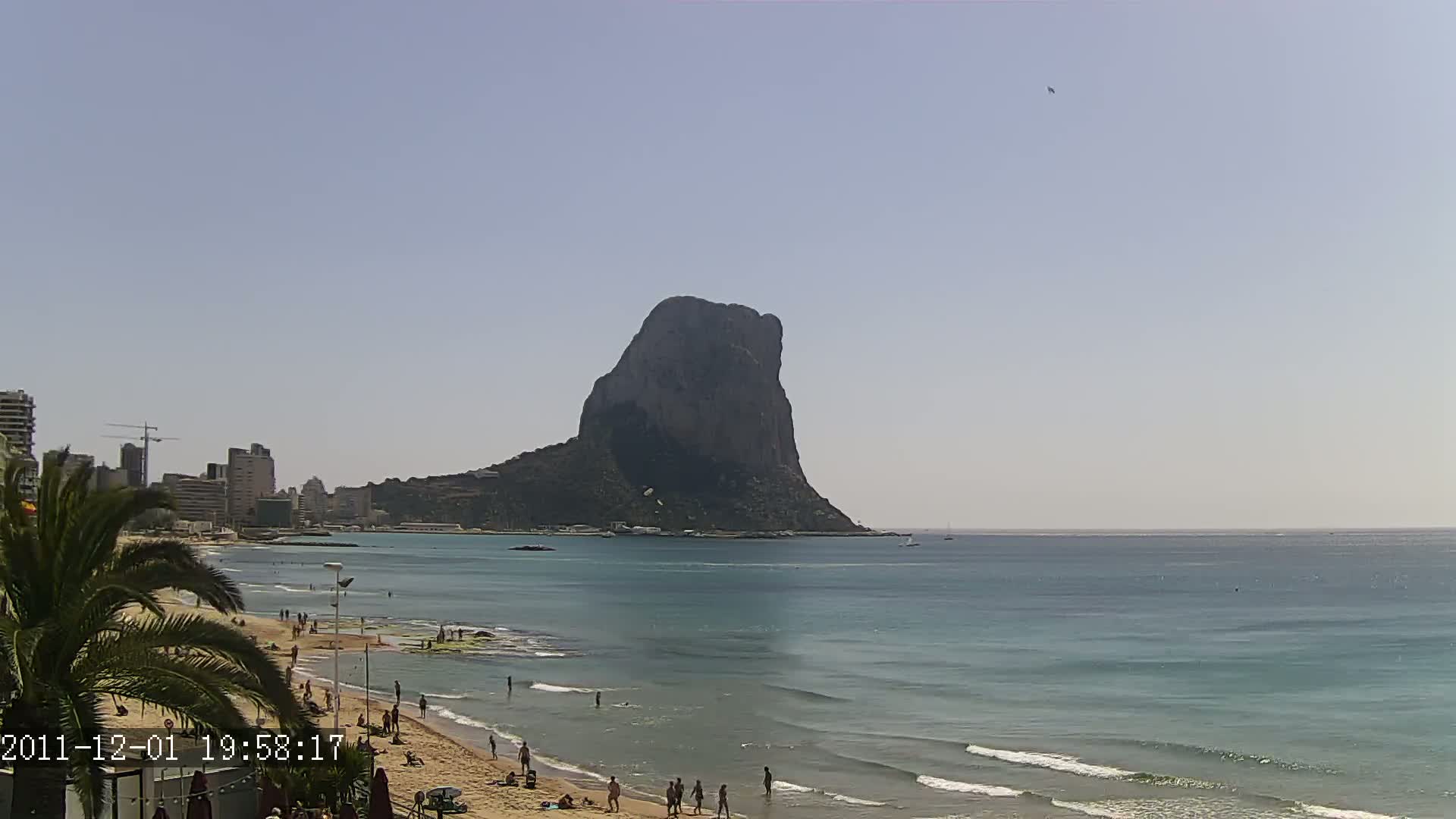 A large, dark rock formation rises from the calm, light blue ocean, with a sandy beach and people visible in the foreground under a clear, sunny sky.