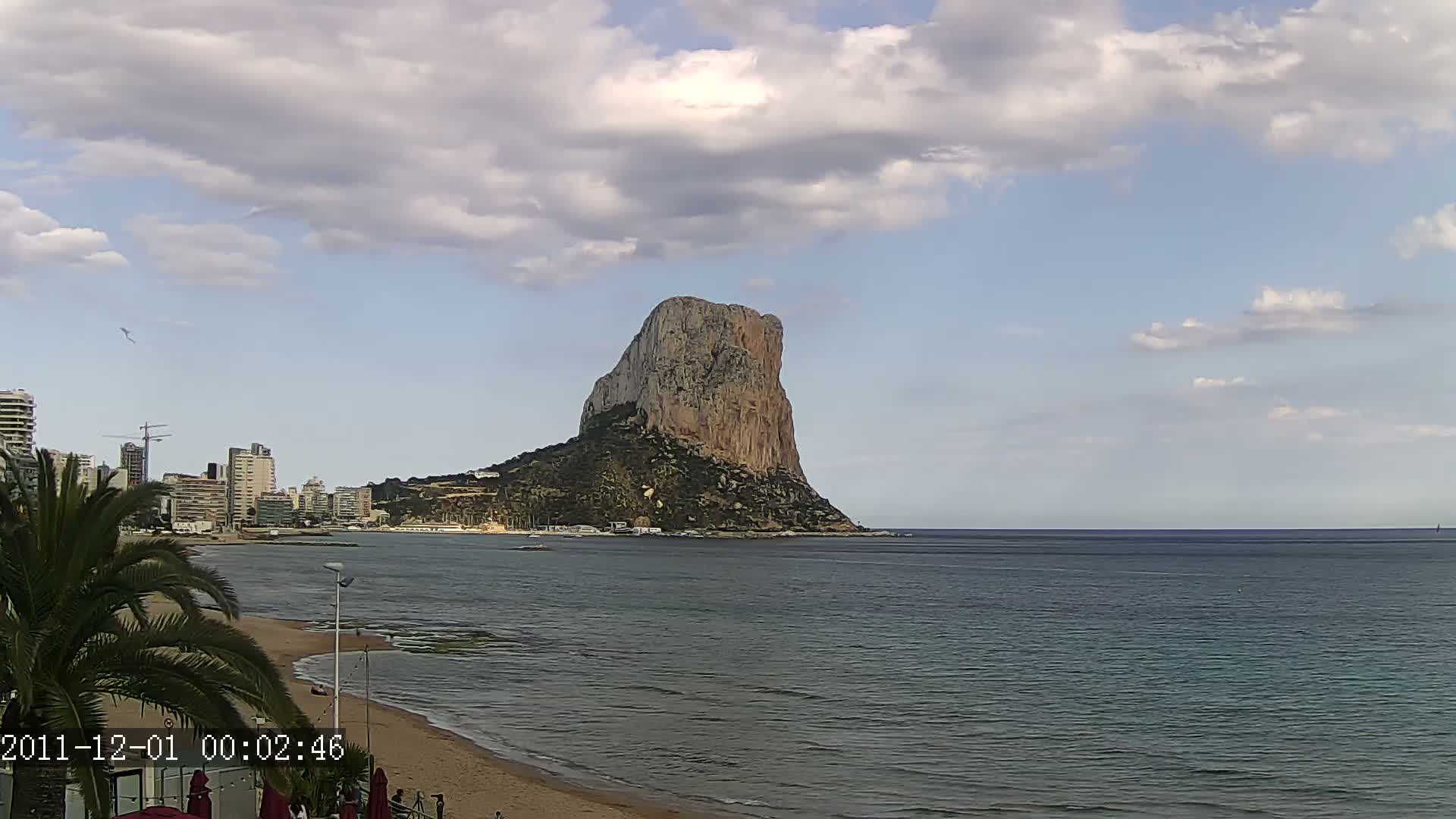 A large, rocky island stands in a calm sea under a partly cloudy sky, with a sandy beach and buildings visible in the foreground.