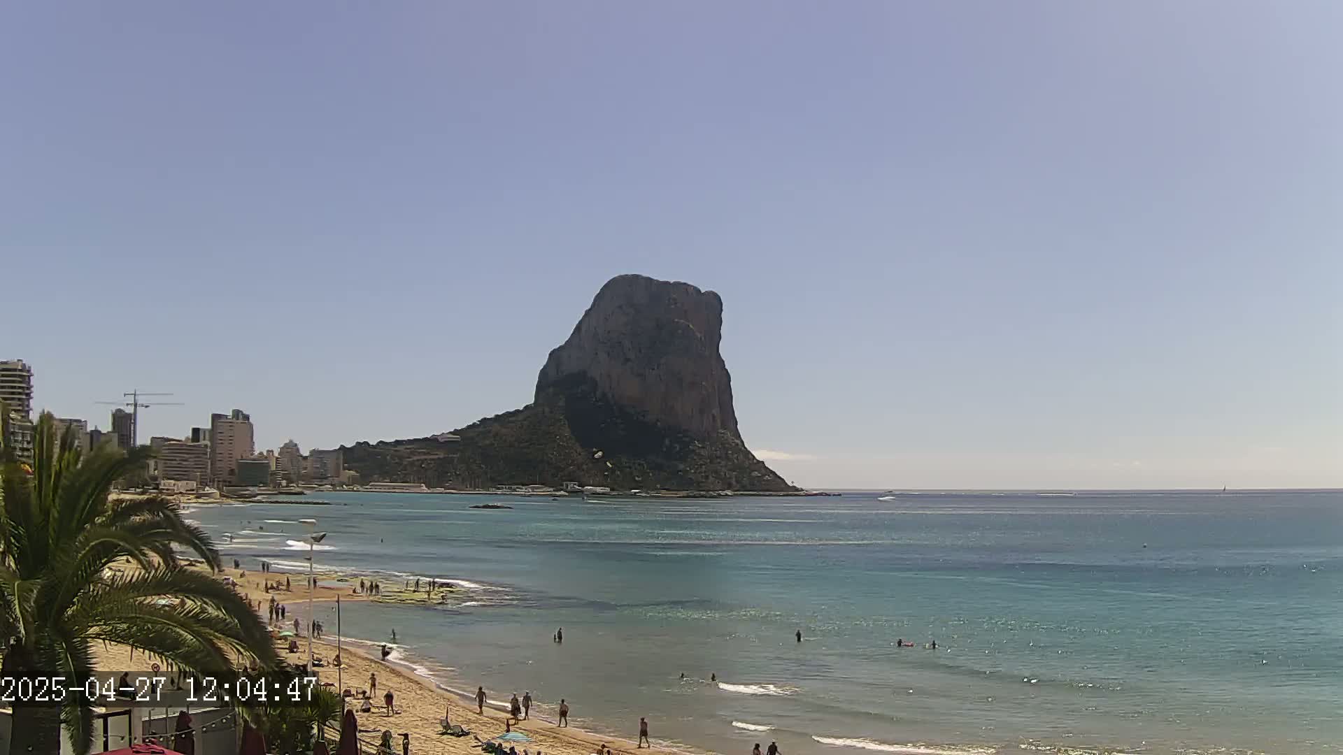 A sandy beach with people swimming and sunbathing, a cityscape in the background, and a large, rocky mountain rising from the sea under a clear, sunny sky.