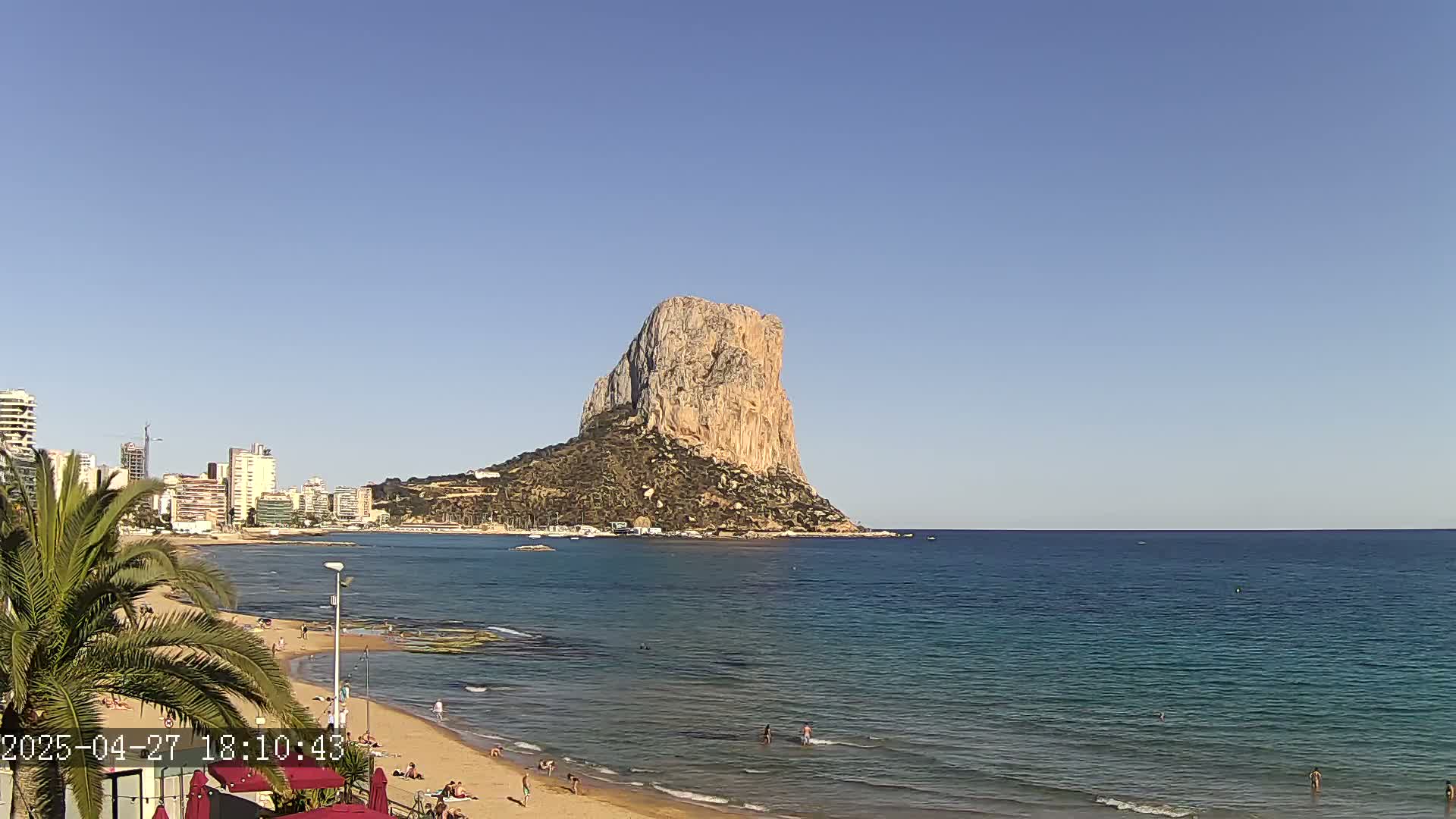 A large, tan rock formation rises from the sea, visible from a sunny beach with people swimming and sunbathing, and a cityscape in the background.