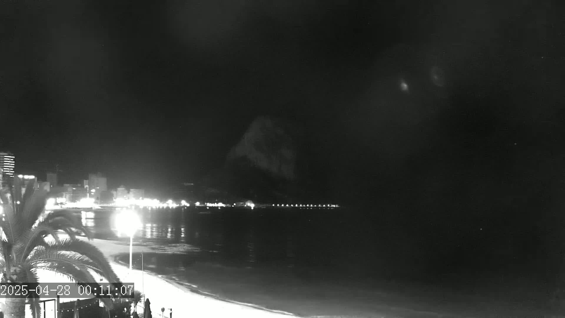 A nighttime view of a beach with city lights and a dark mountain in the background under a clear sky.