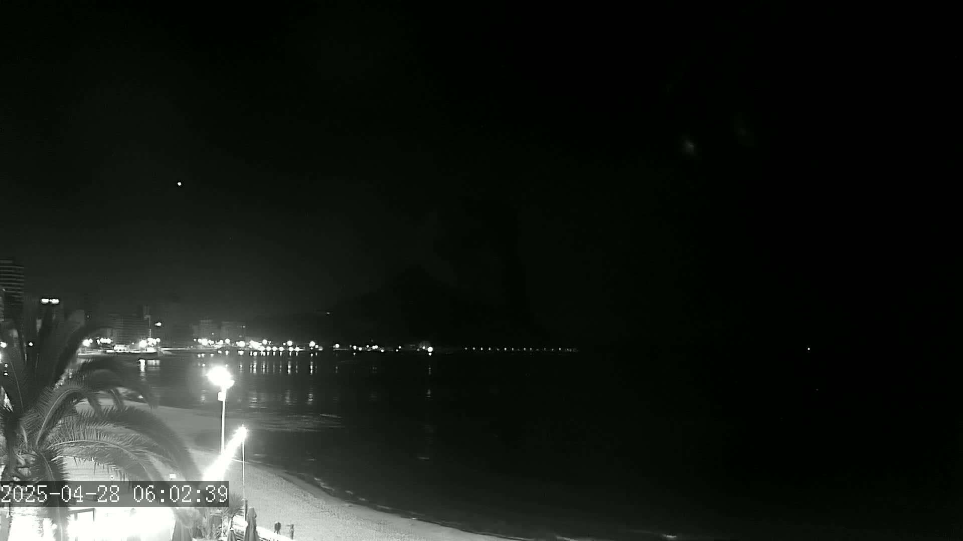 A nighttime view of a beach with city lights across the water under a clear, dark sky.