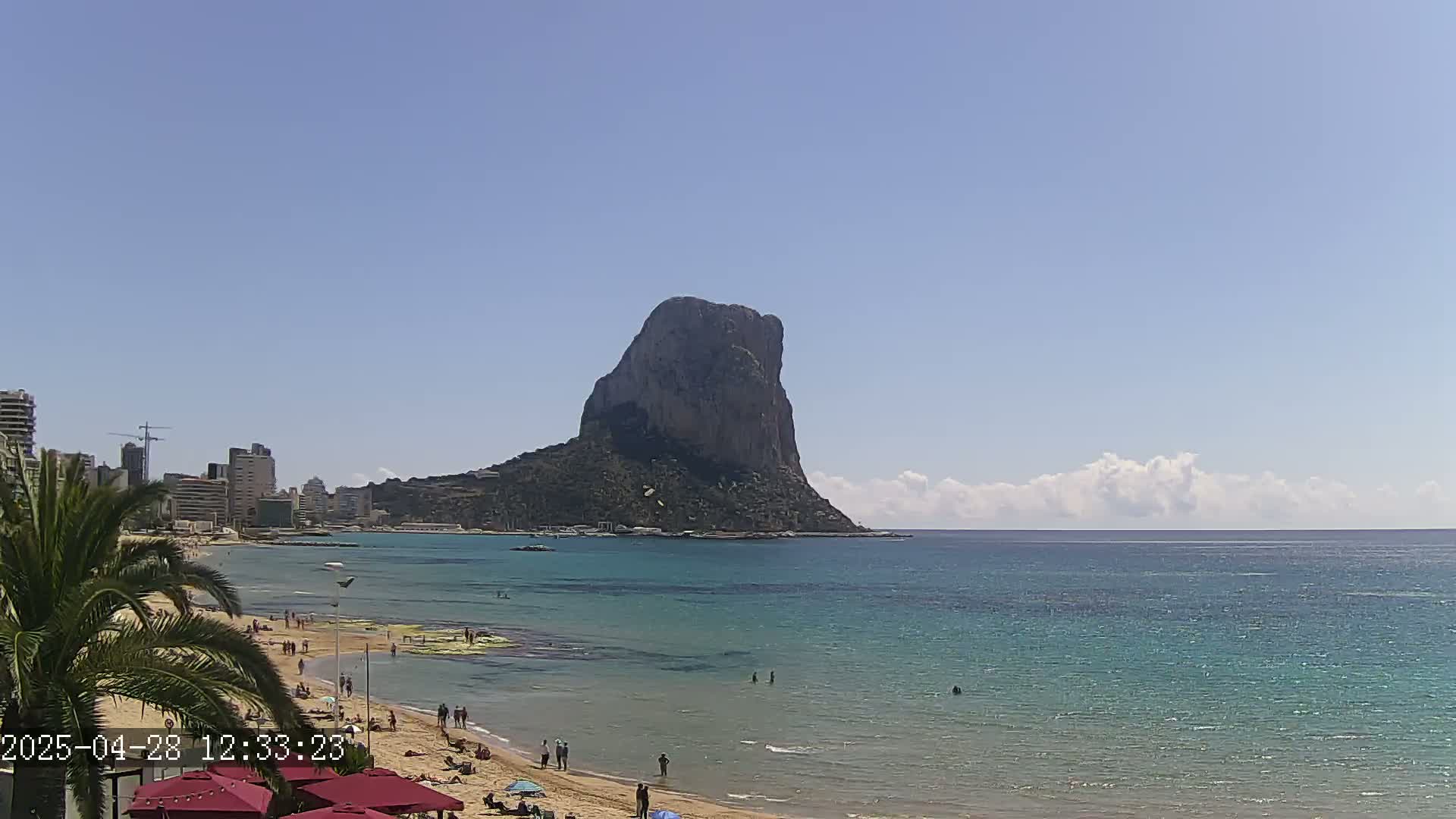 A sandy beach with people swimming and sunbathing stretches along a calm, turquoise sea, with a large, imposing rock formation visible in the distance under a mostly sunny sky with a few clouds.